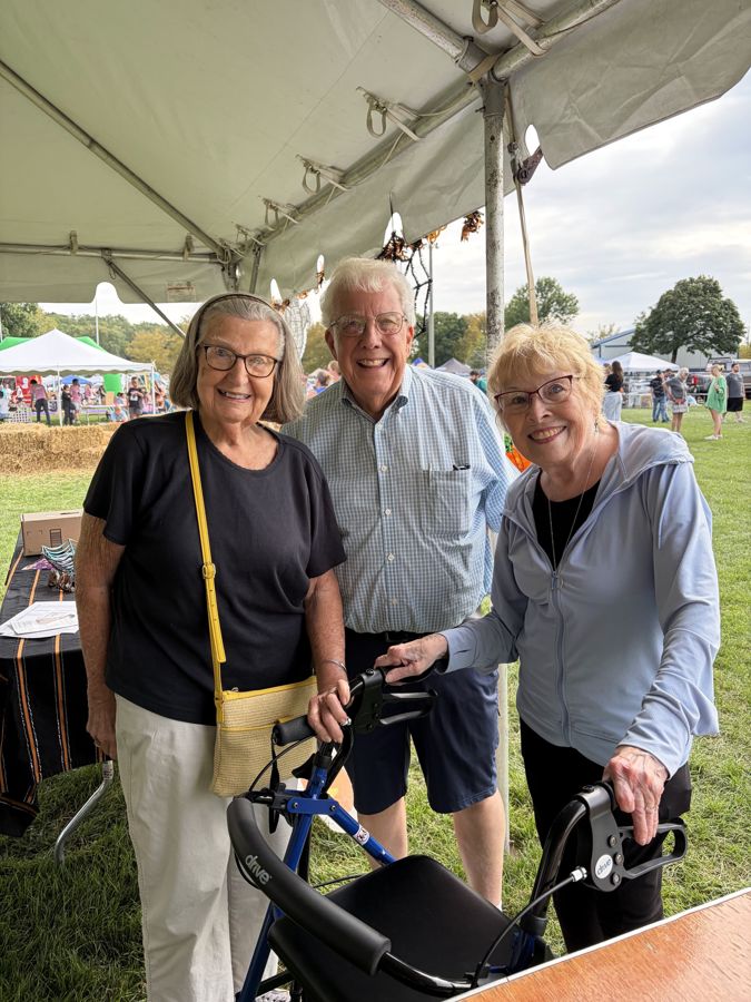 Three longtime neighbors smiling together under a tent at Community Day 2026